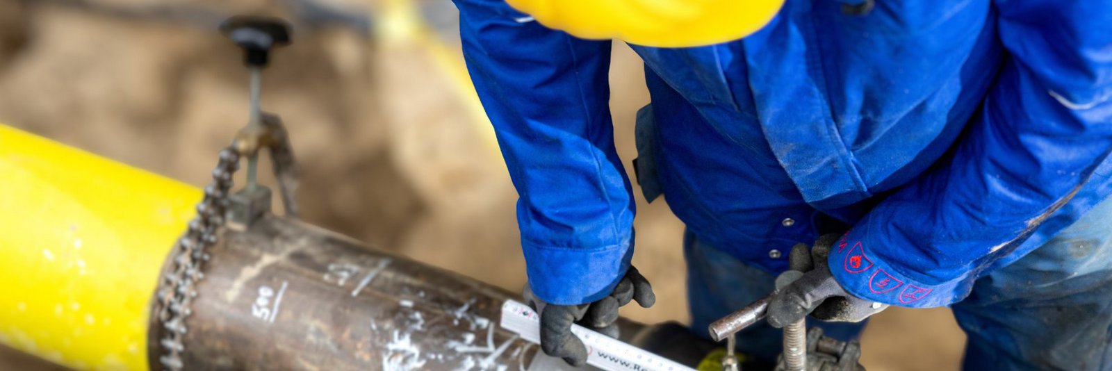 Eine gelbe Gasleitung in einer Baugrube von oben fotografiert, ein Mitarbeitender steht in Arbeitskleidung daneben und arbeitet direkt an der Leitung.