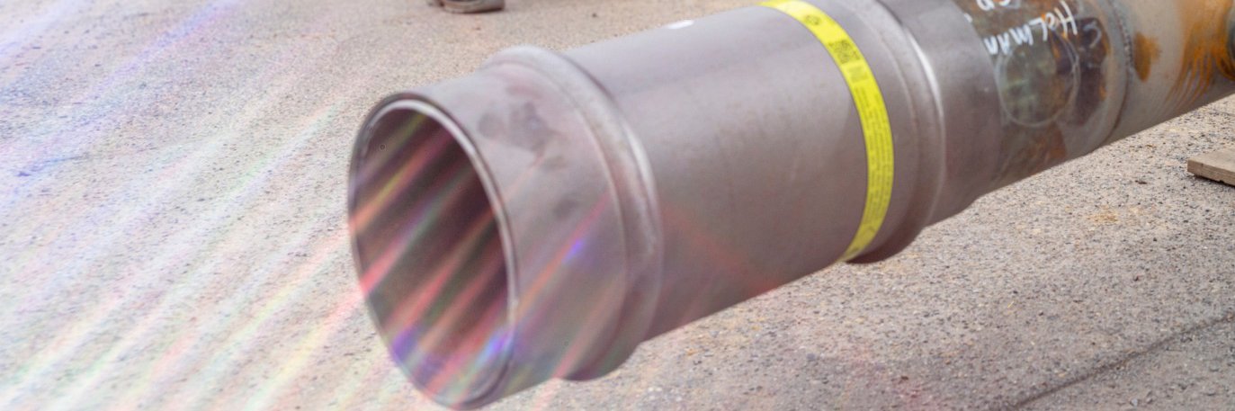 Eine starke Gasleitung liegt im Vordergrund auf einer Straße vor einer Baustelle, dahinter stehen drei Monteure in Sicherheitsbekleidung, die sich unterhalten. 